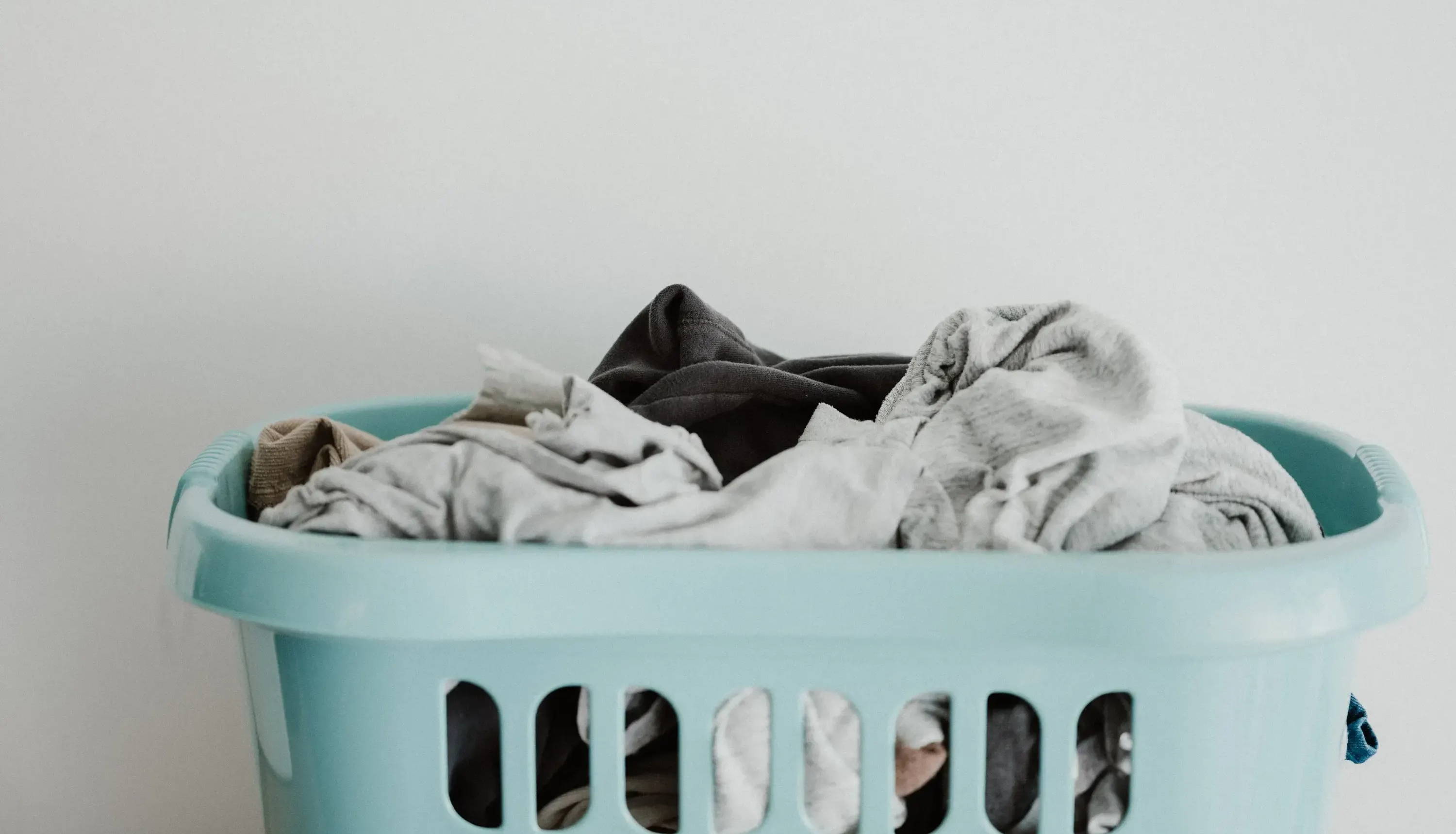 A blue laundry basket sits in front of a white surface filled with dirty clothes. Image by Annie Spratt. 