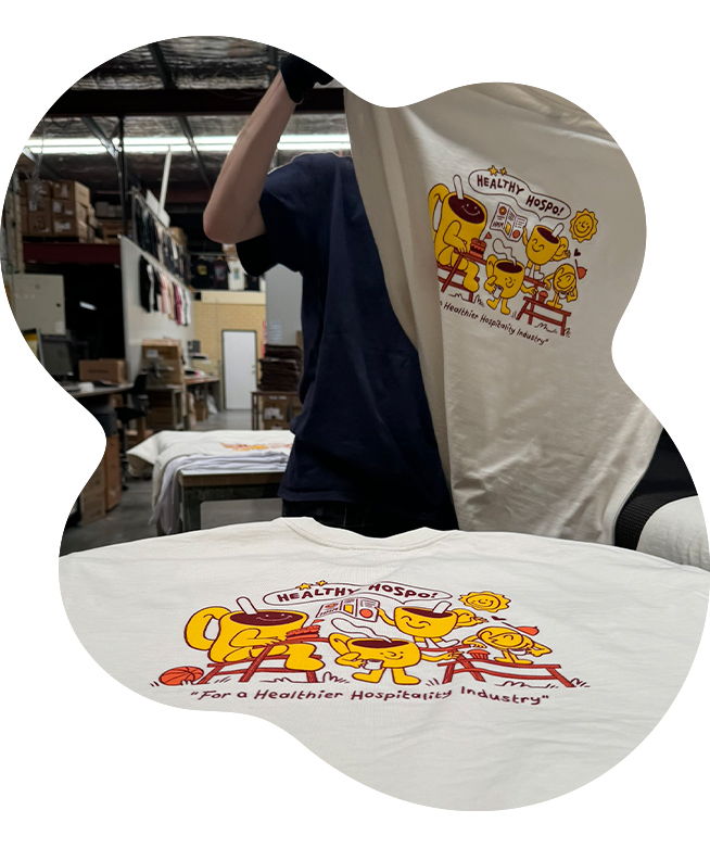 Staff member in a warehouse holding up a cream t-shirt screen printed with the Healthy Hospo colourful cartoon character graphic, with a second printed t-shirt laid flat in the foreground