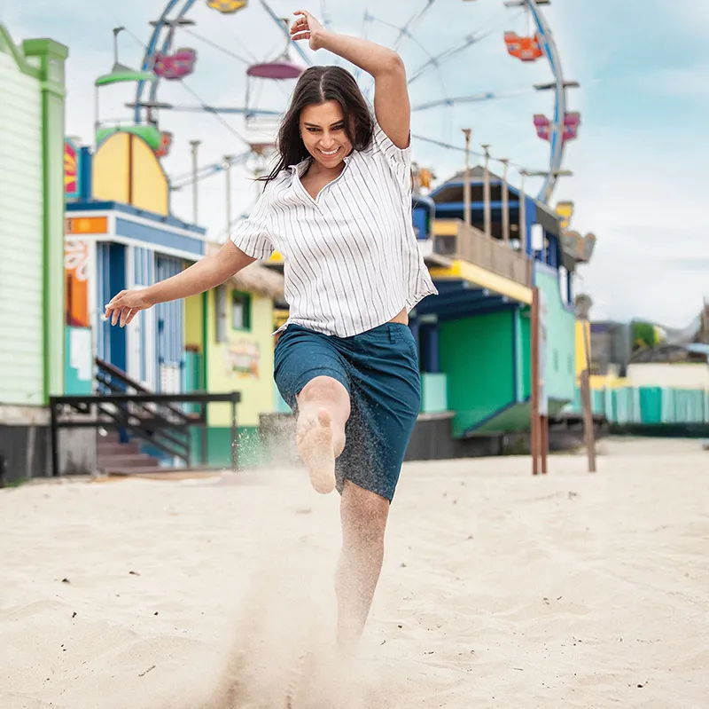 Person kicking up sand on a beach, with colorful buildings and a ferris wheel.