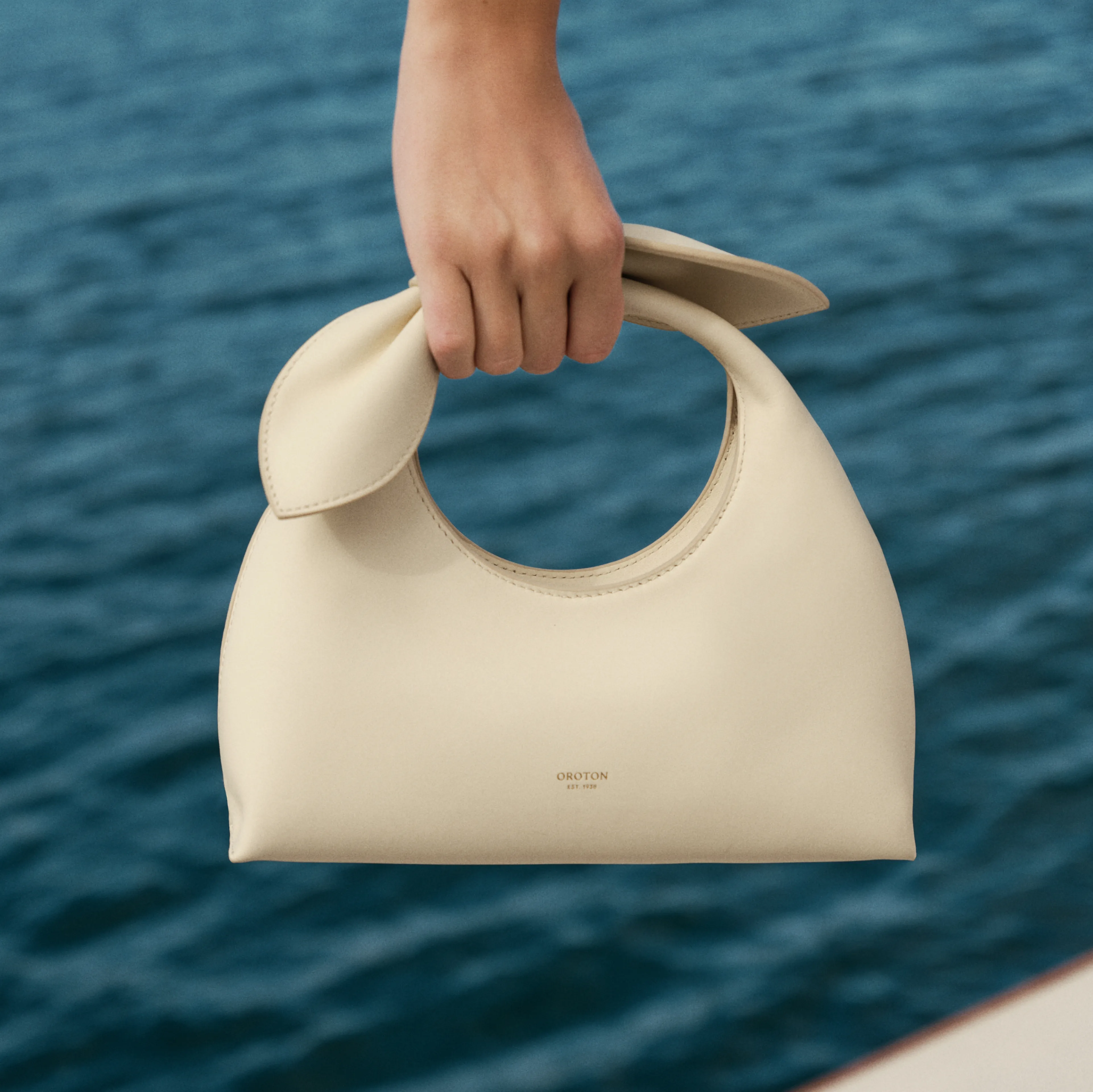 A hand holding a cream-colored handbag against a water background.