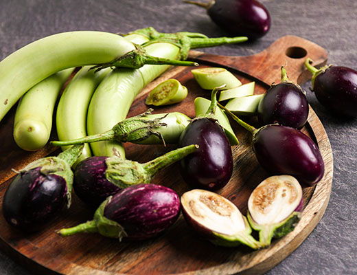 Various eggplants and green bananas on a wooden cutting board.