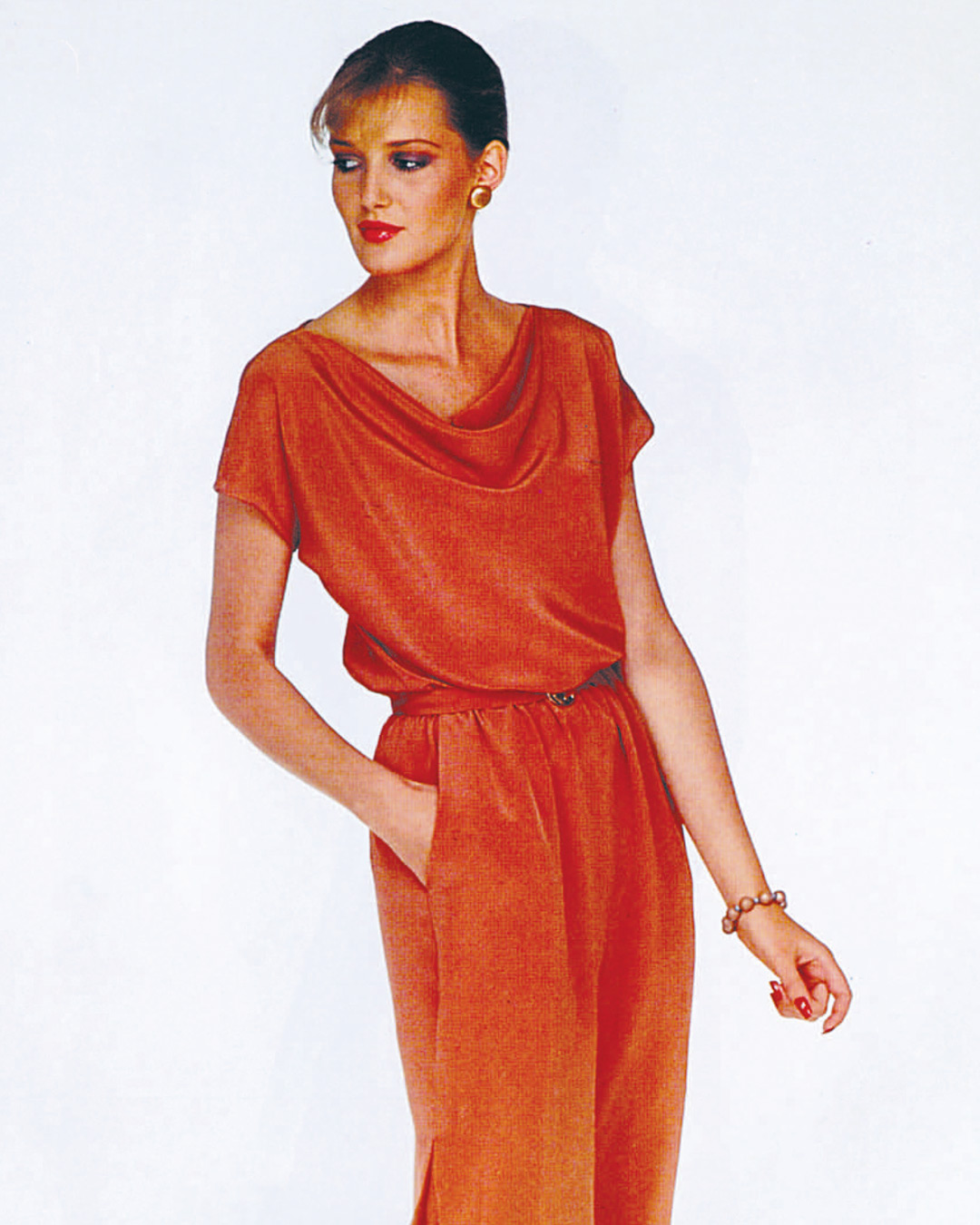 Model wearing an orange dress with a draped neckline and hands in pockets.
