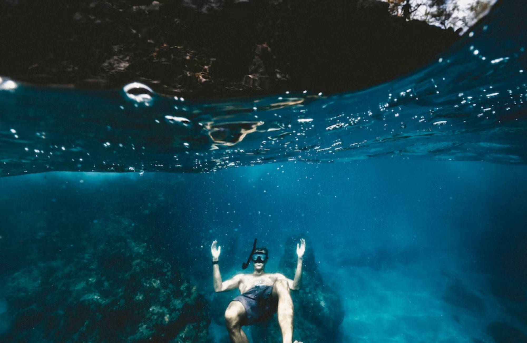 man floating below the surface in clear, dark blue water, wearing a snorkel mask and swim trunks.
