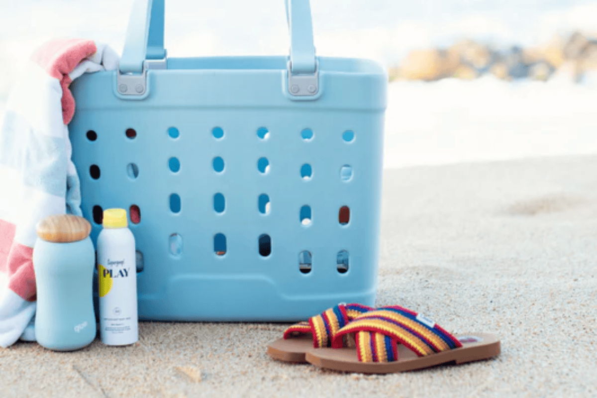 Blue beach tote on sand with towels, sunscreen, a water bottle, and striped sandals near the shoreline.