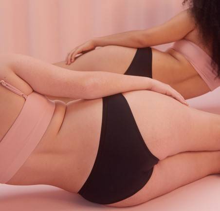 Fotografia de duas mulheres deitadas de lado, de costas para a foto, utilizando top e calcinha absorvente