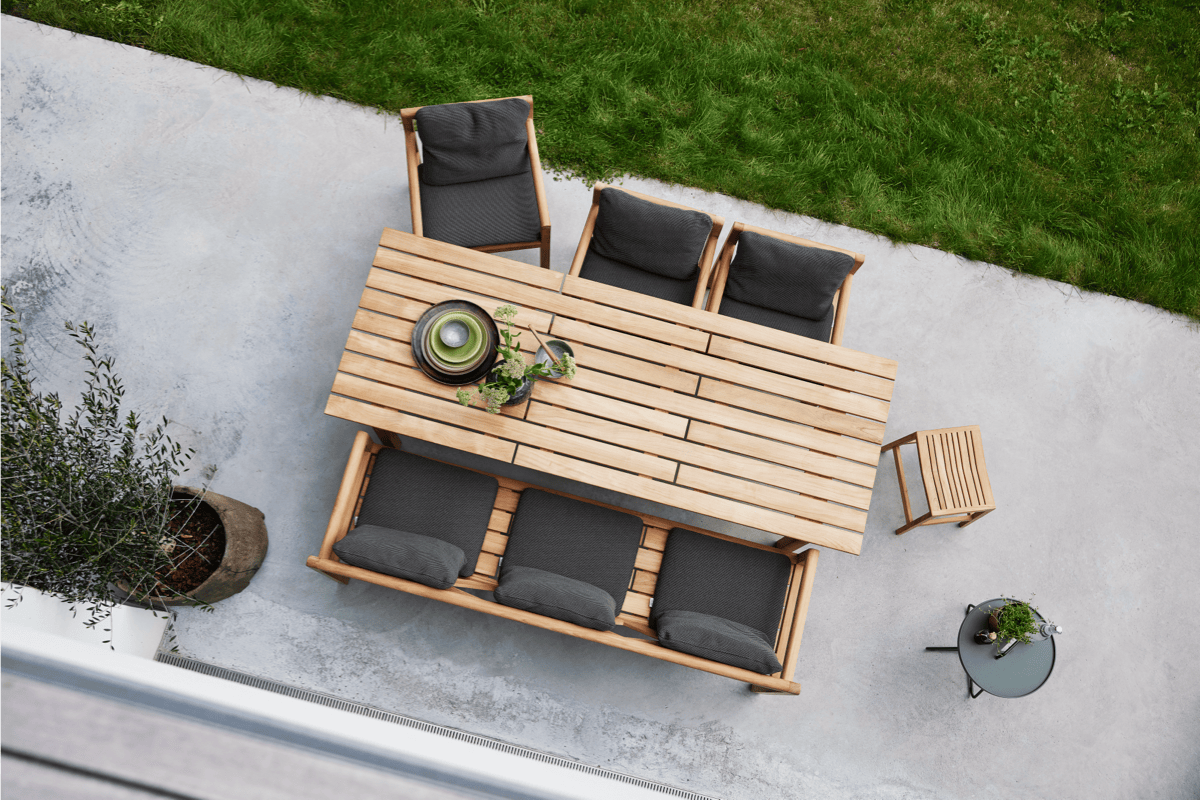 A teak dining table and chairs with dark grey cushions on a concrete patio with green grass nearby.