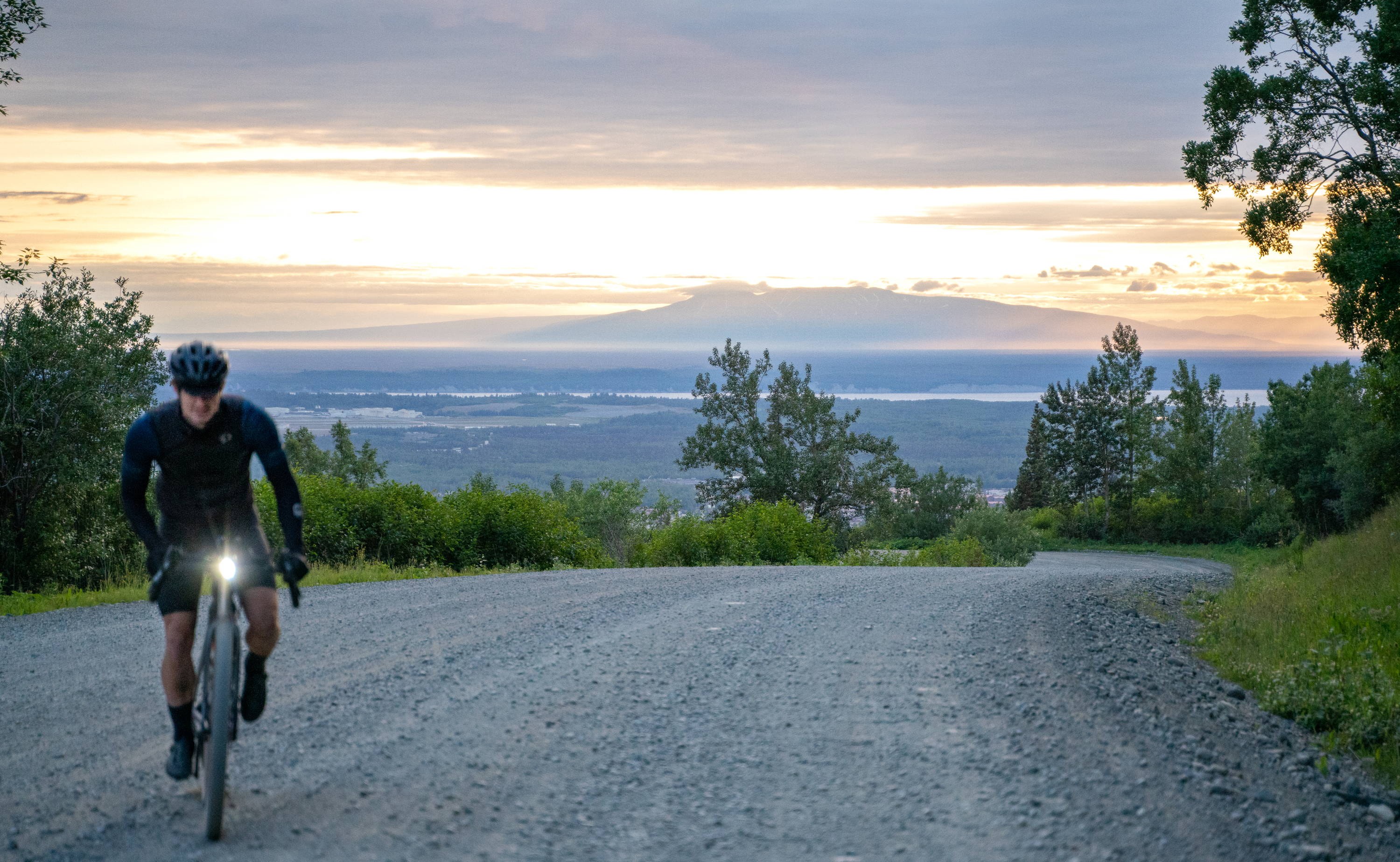 Dylan Morton charging up a gravel hill on his Otso Cycles bike. The gravel road curves off behind him, revealing a mountainous sunset.