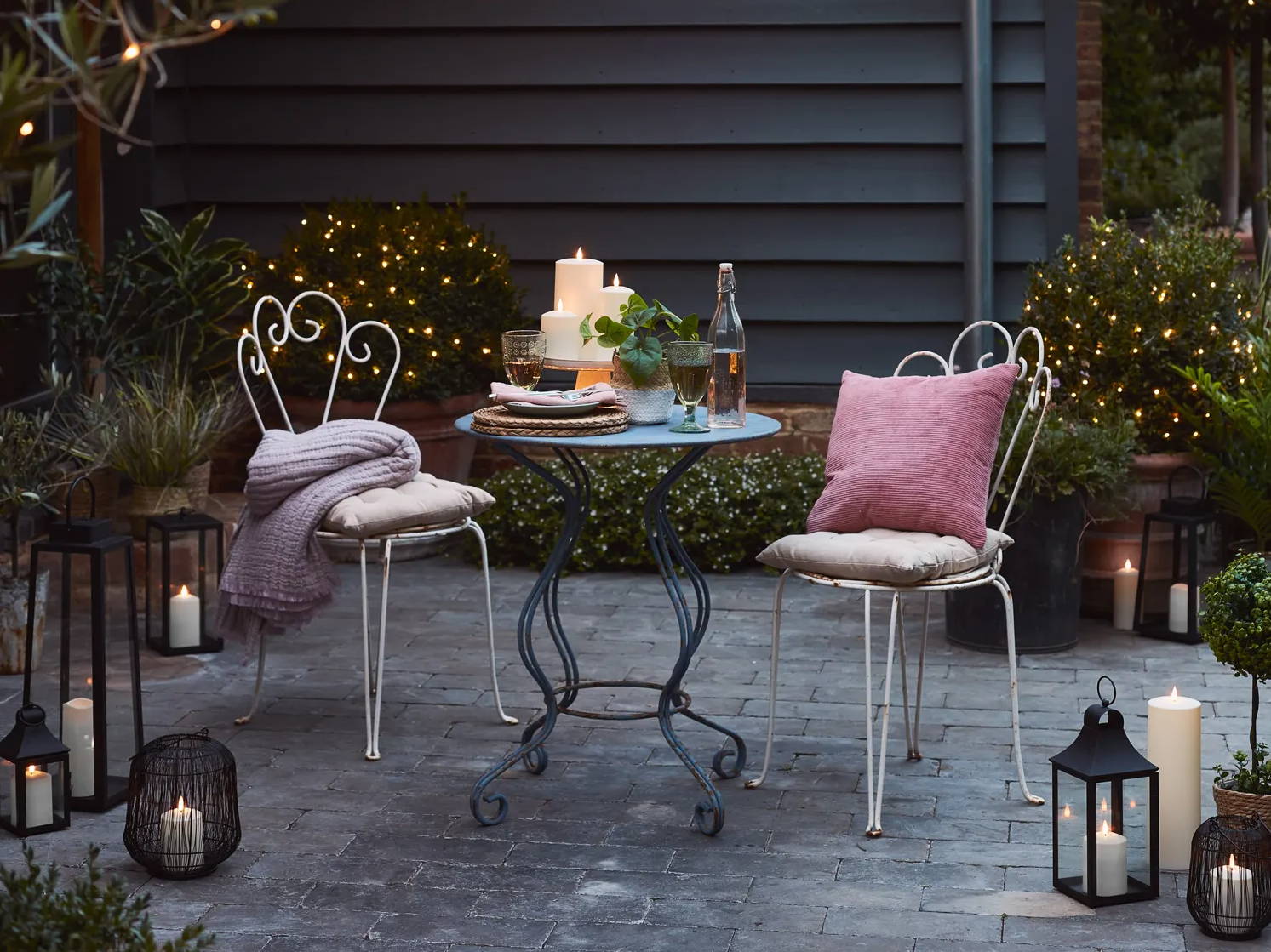 Eine Sitzecke auf der Terrasse mit Laternen, LED-Kerzen, einer Solar-Pendelleuchte und warmweißen Lichterketten.