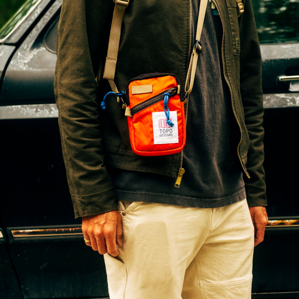 A person wearing a black jacket and beige pants, holding an orange bag.