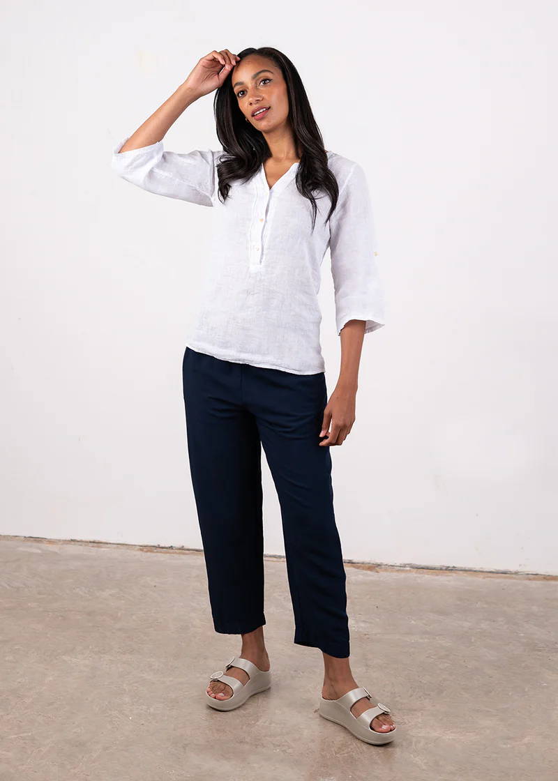 A model wearing a white linen semi sheer top with long sleeves and button down neckline with black cropped trousers and off white platform slides