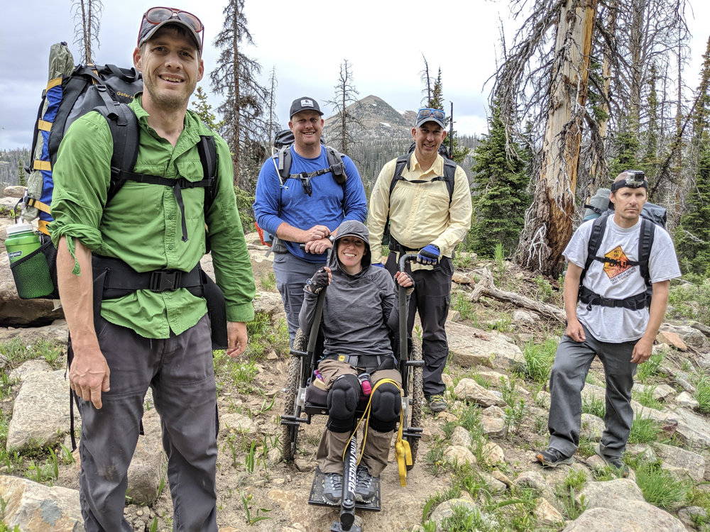 Group hiking, including one using GRIT Freedom Chair all-terrain wheelchair,  on rocky grass and dirt area among trees