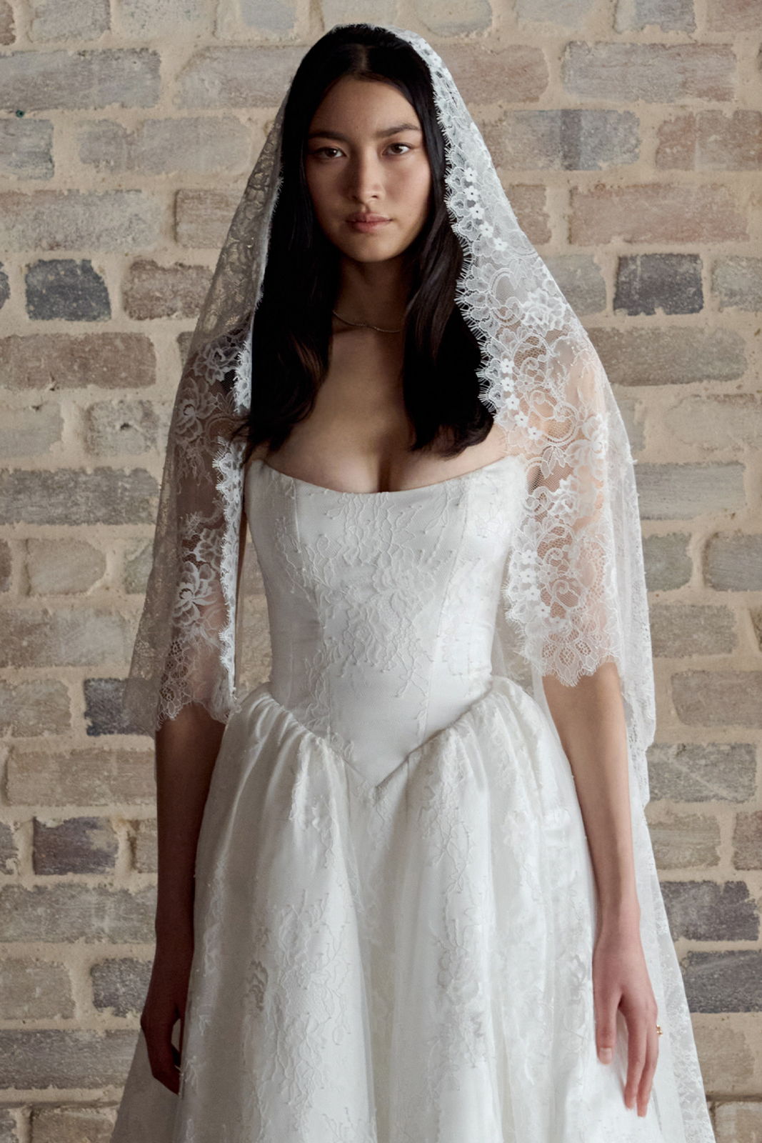Bride in a white wedding dress with a lace veil against a brick wall.