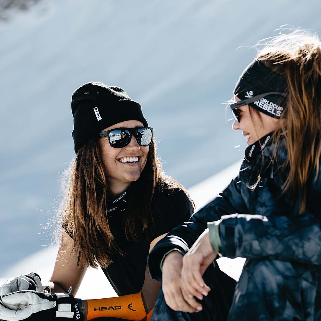 Two women sit and talk on a snowy hill; one of them is wearing an eSmartr smart compression sleeve from HEAD™.