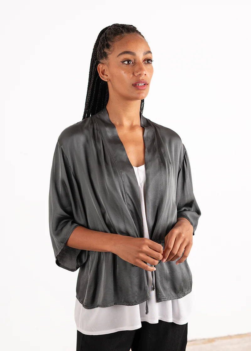 A model wearing a dark grey satin jacket with kimono style sleeves over a white top