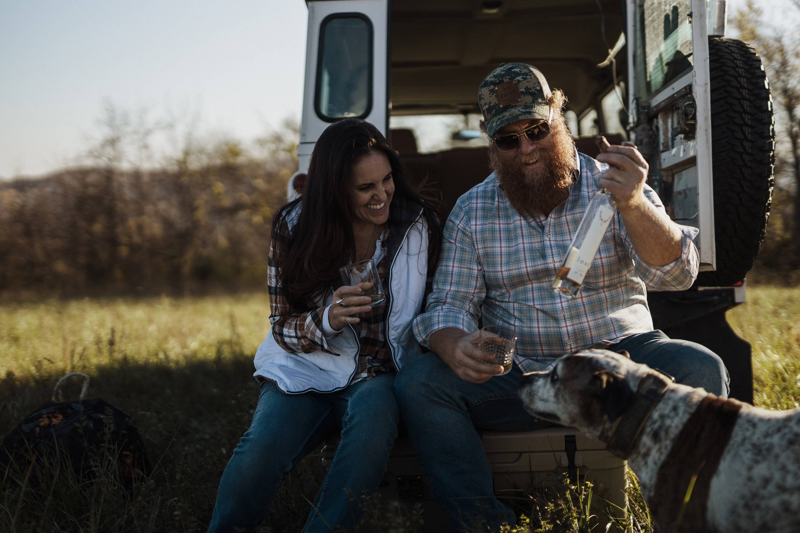 A couple (man and woman) sitting on the bumper of a vintage vehicle looking at a bottle of Lost Whiskey.