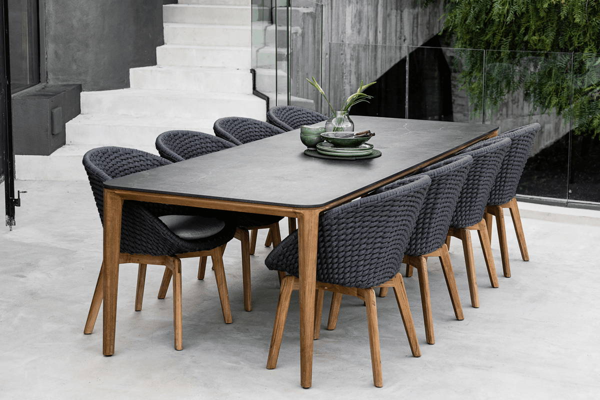 Rectangular outdoor dining table with black stone top and teak legs set with 8 charcoal grey woven chairs.