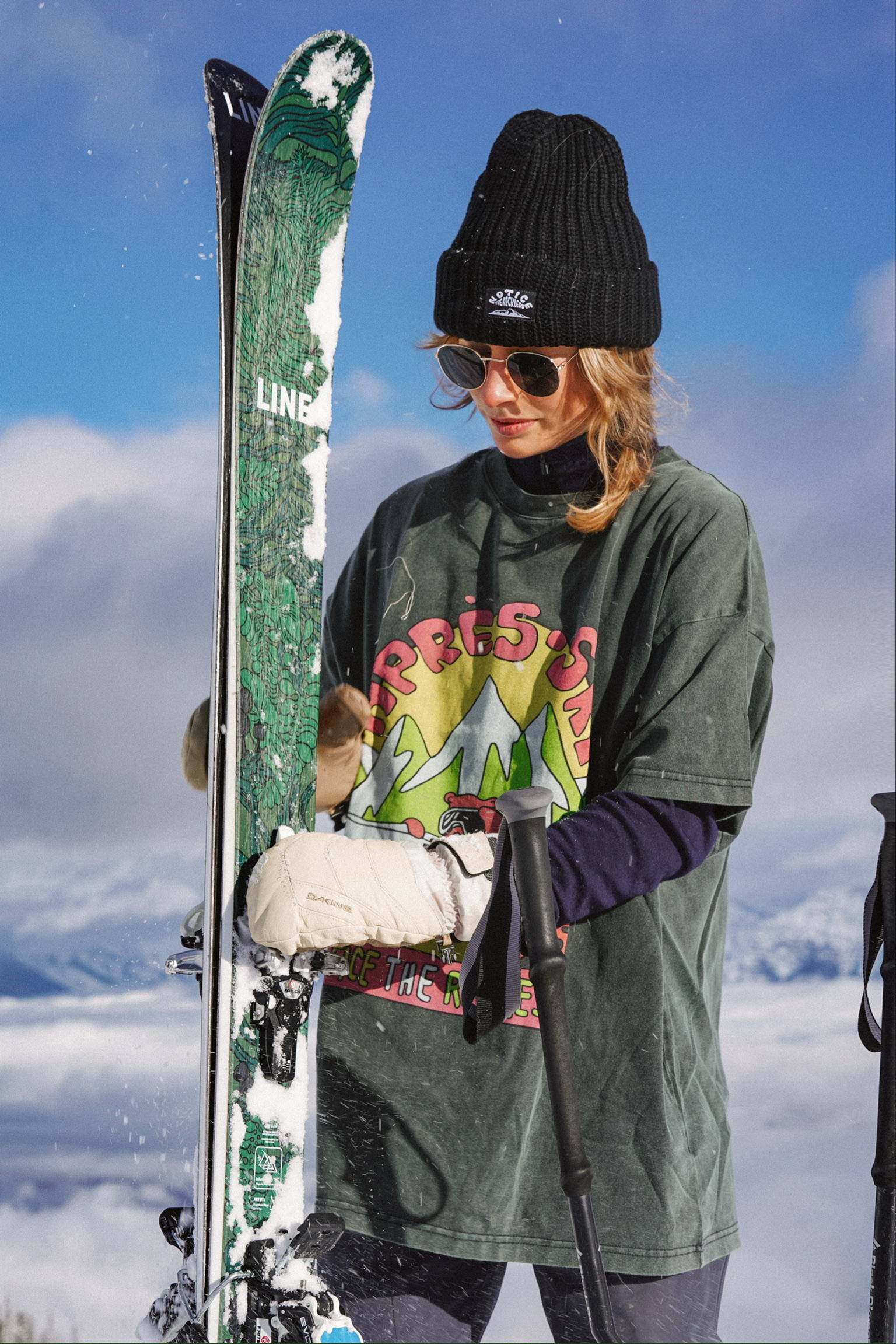 A woman holding her ski on top of a mountain wearing an oversized t-shirt and a black beanie