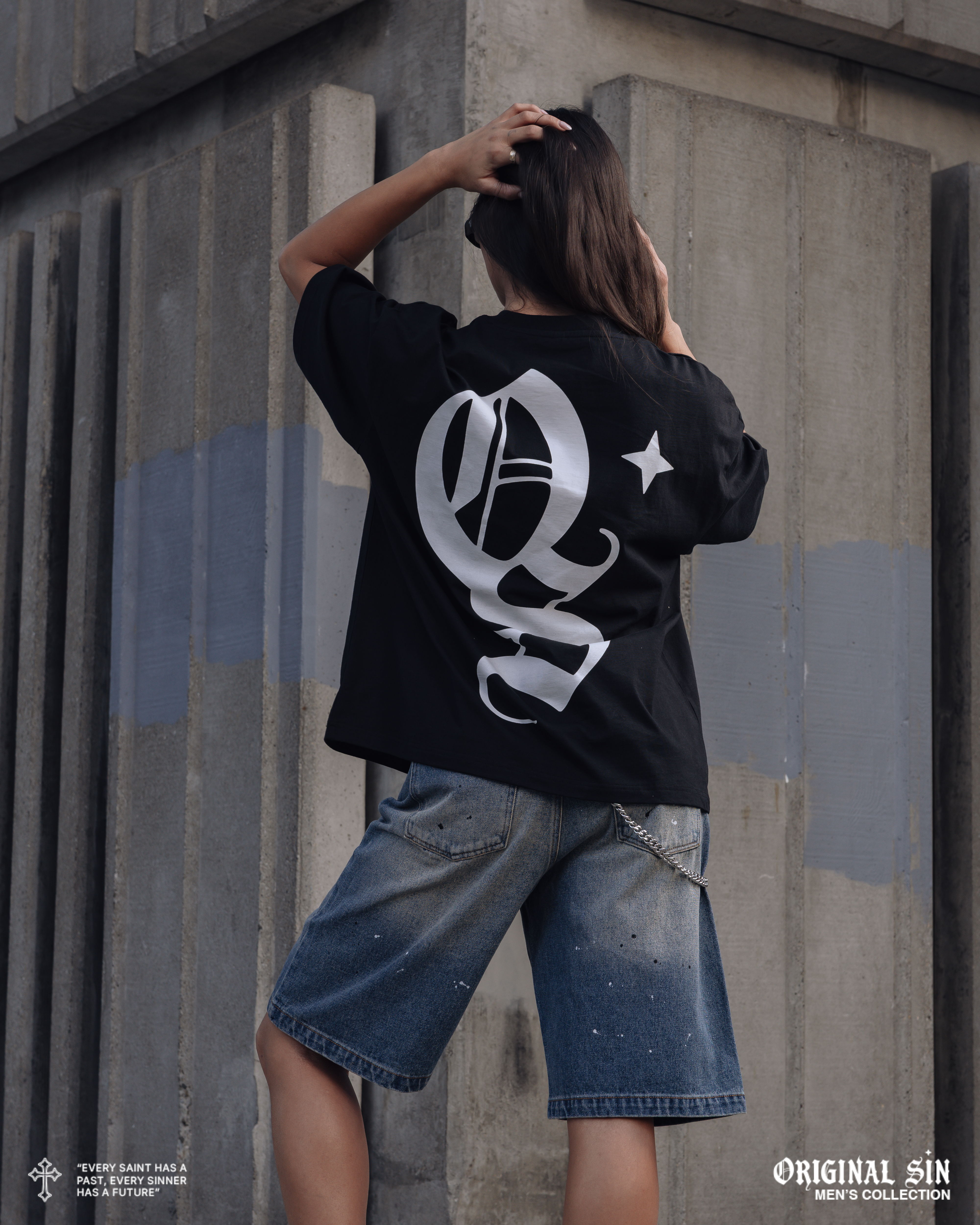 Person wearing a black shirt and denim shorts, posing against a wall.