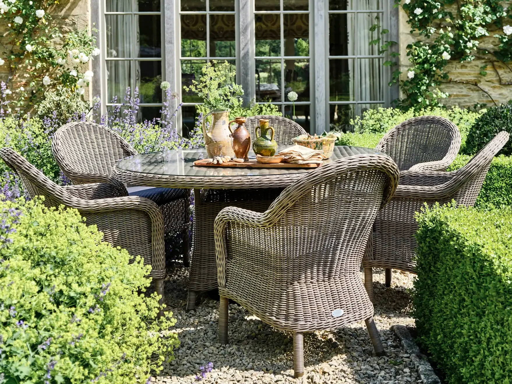Garden rattan dining set with 6 dining armchairs and a round dining table surrounded by hedges.