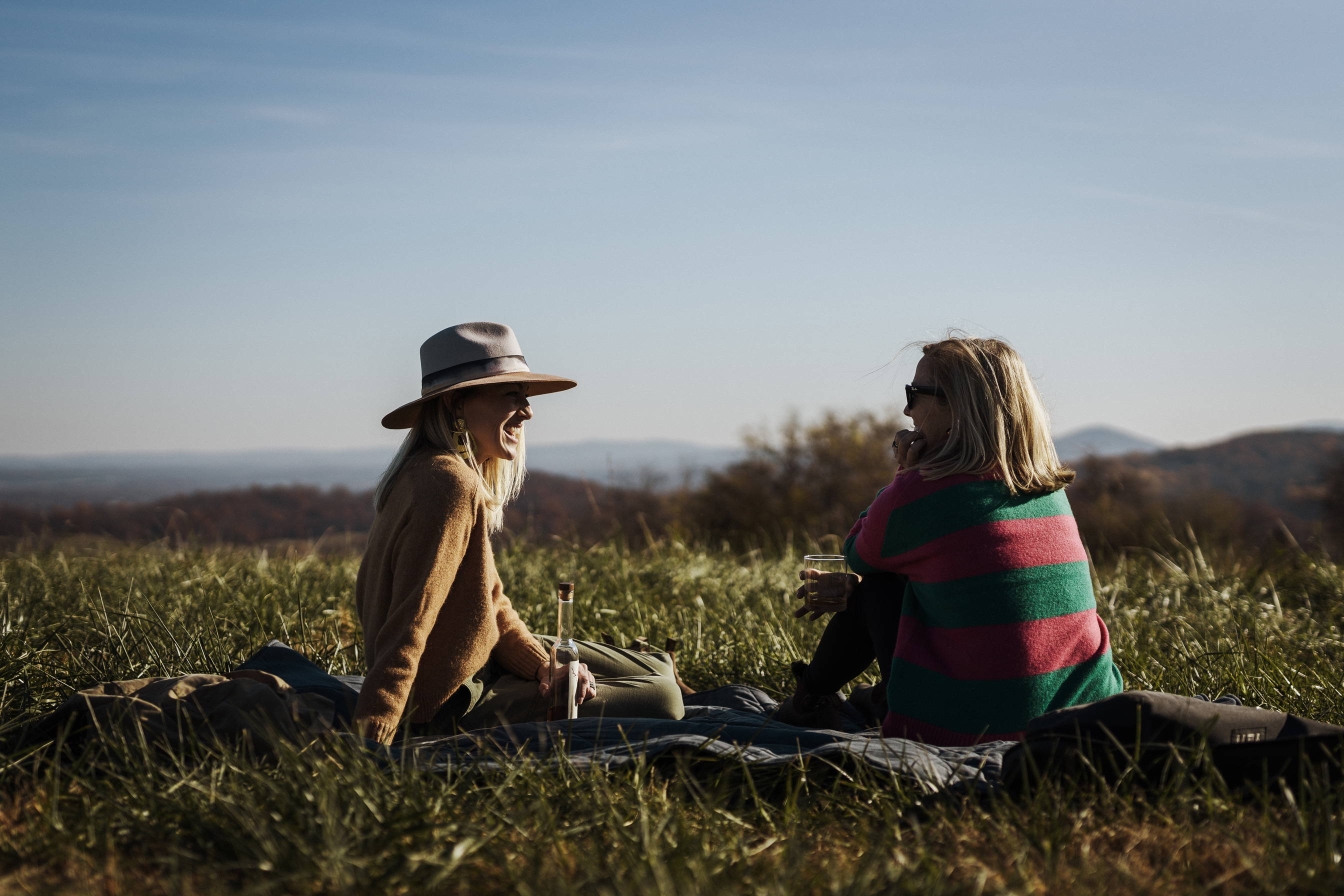 Two women sitting in a field on a picnic blanket with a view of the mountains.