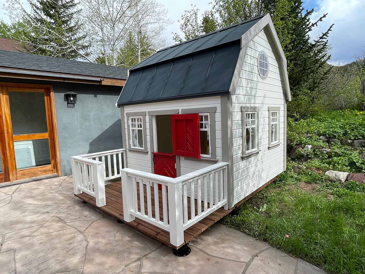 Kids Fully Finished Playhouse in white color with red dutch door and wooden terrace in a backyard by WholeWoodPlayhouses