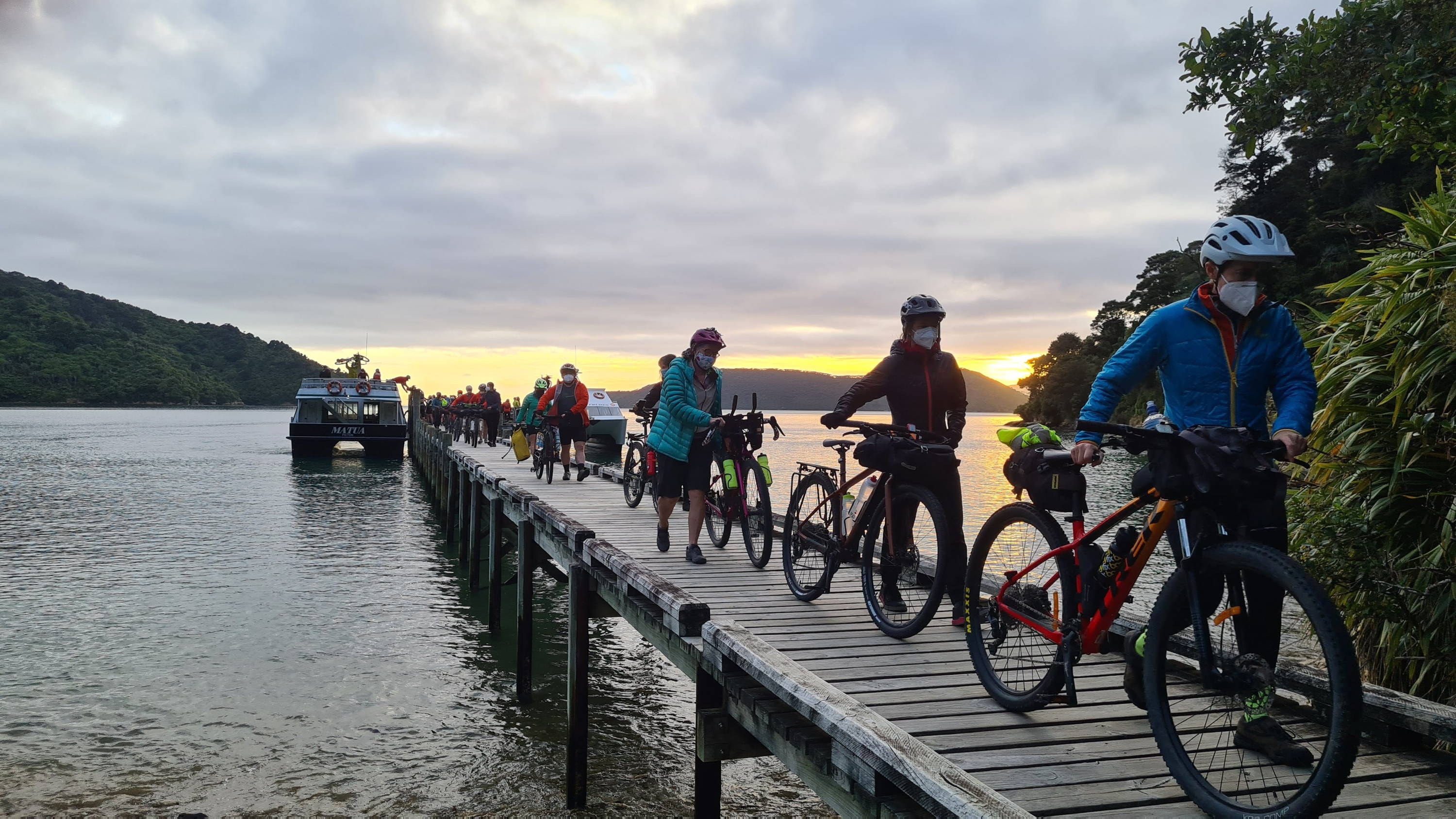 A line of cyclists walk their bikes across a boat dock toward the shore in early dawn.