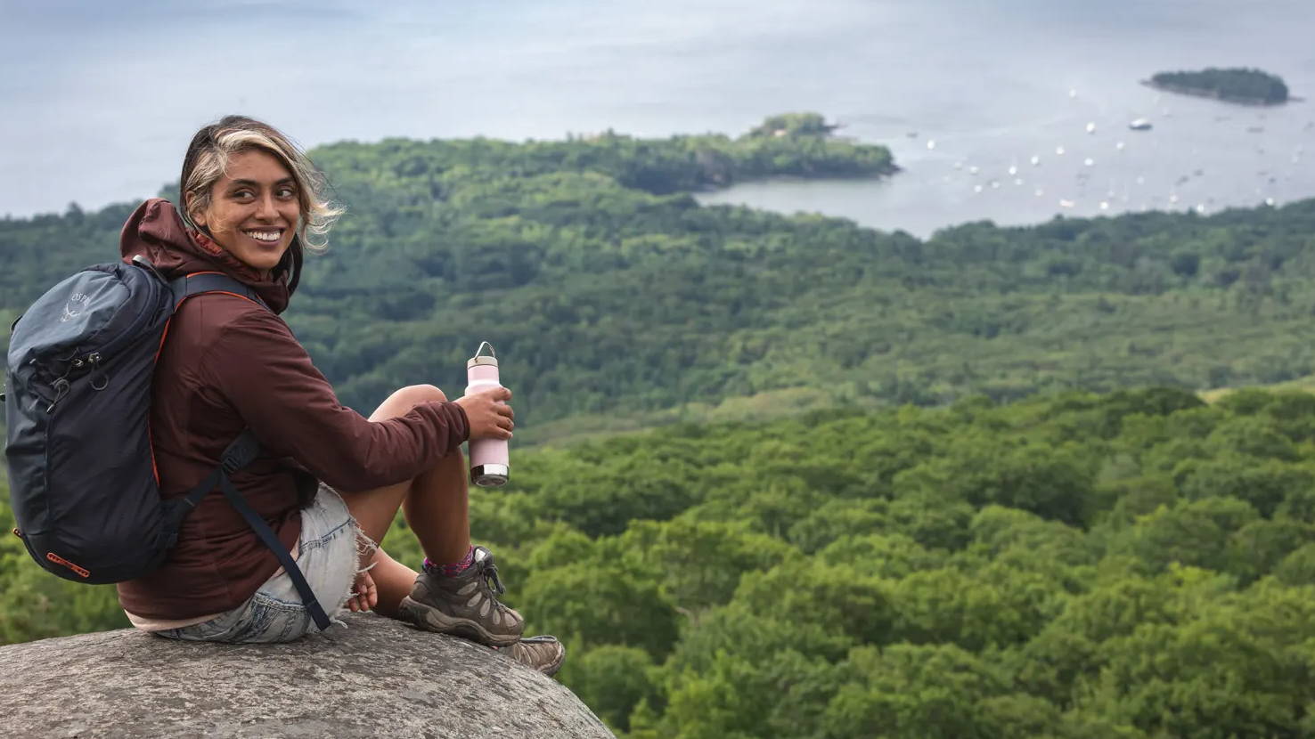 female bikepacker sitting on the top of a rock overlooking green trees and the ocean with a klean kanteen classic insulated bottle