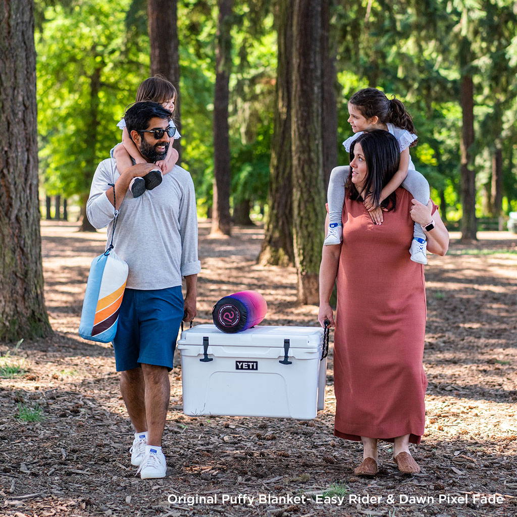 Two people and two kids holding rumpl blankets and a cooler in a park
