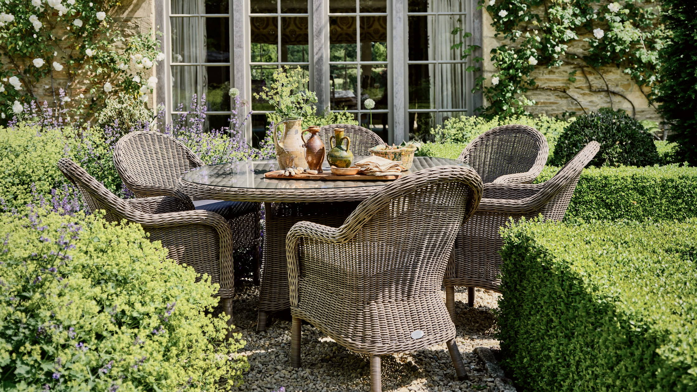 A beautiful rattan dining set surrounded by hedges and ivy sits on a shingled pathway in front of a large window.