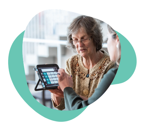 Older woman using a speech-generating device with a younger person assisting her.