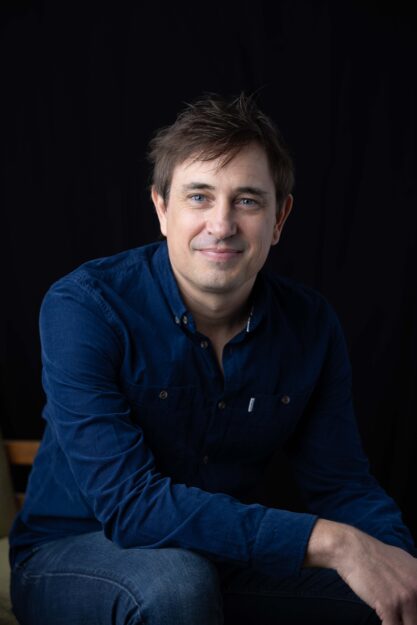 Smiling man in a blue shirt against a dark background.