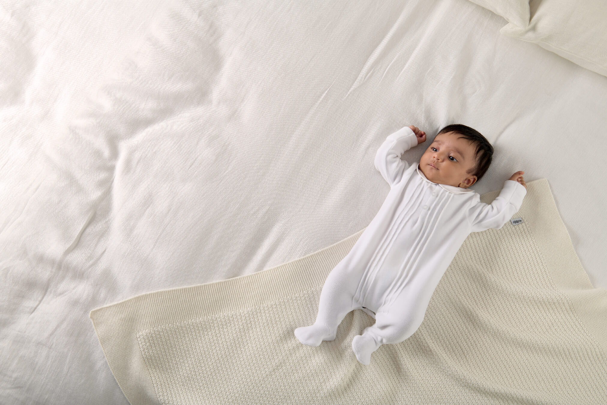 Baby in white onesie lying on a bed with white bedding.