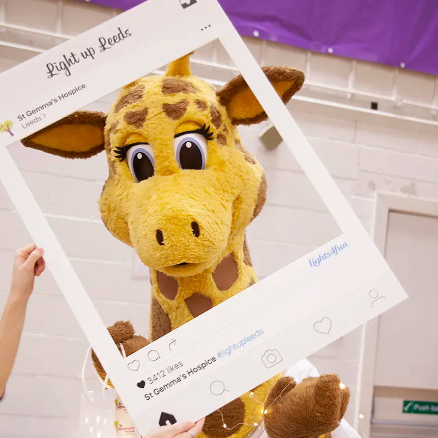 A giraffe mascot posting with an Instagram post sign wrapped in fairy lights. 