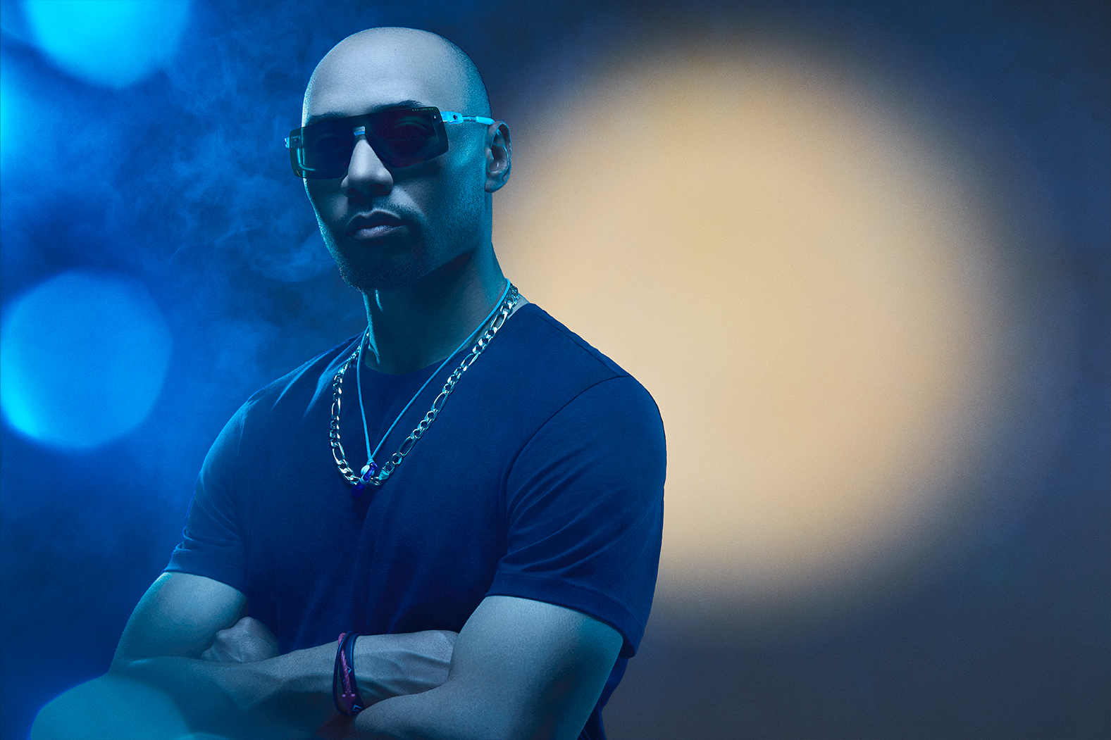 A man with sunglasses poses confidently against a colorful, blurred background.