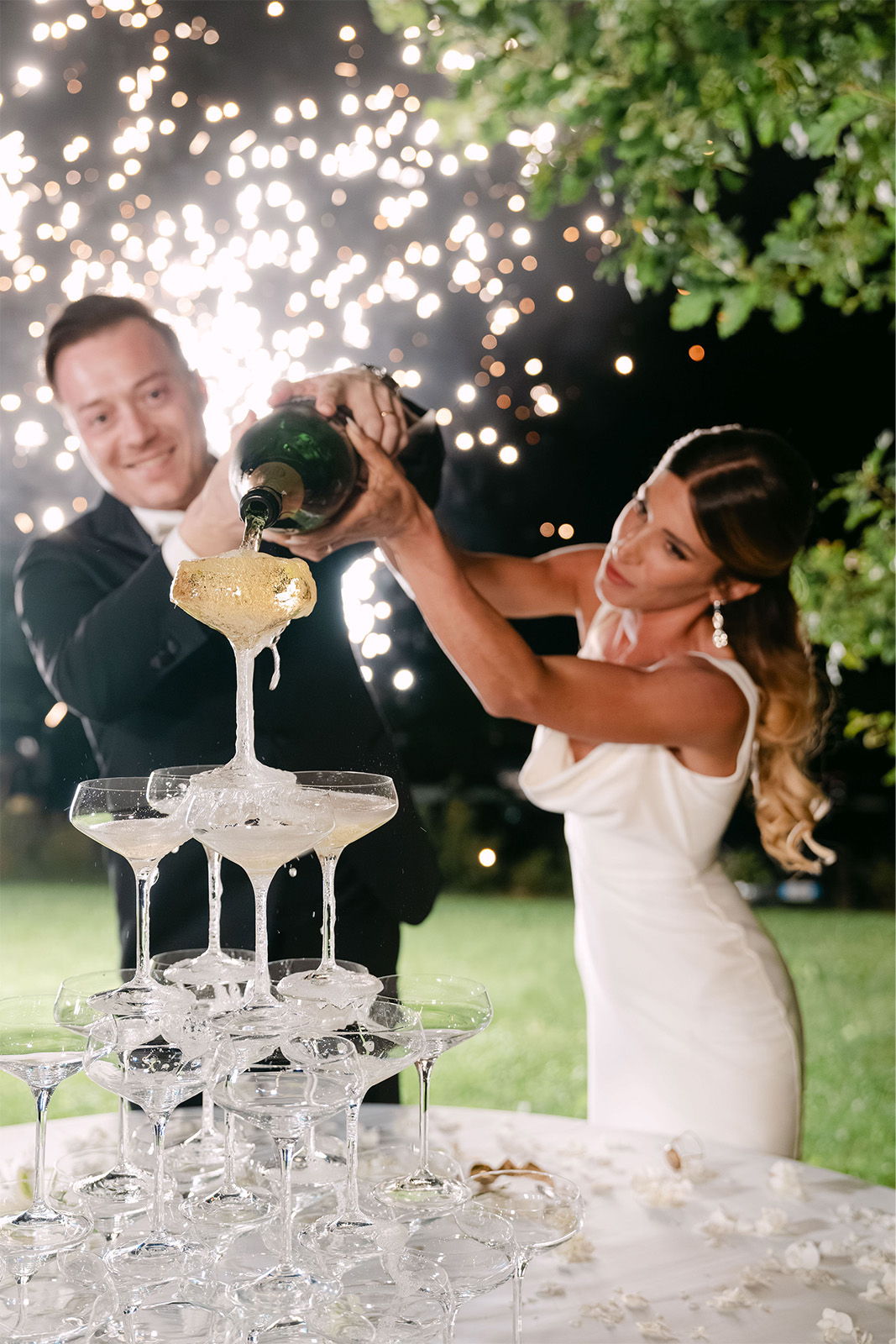 Couple pouring champagne into a glass tower with fireworks in the background.
