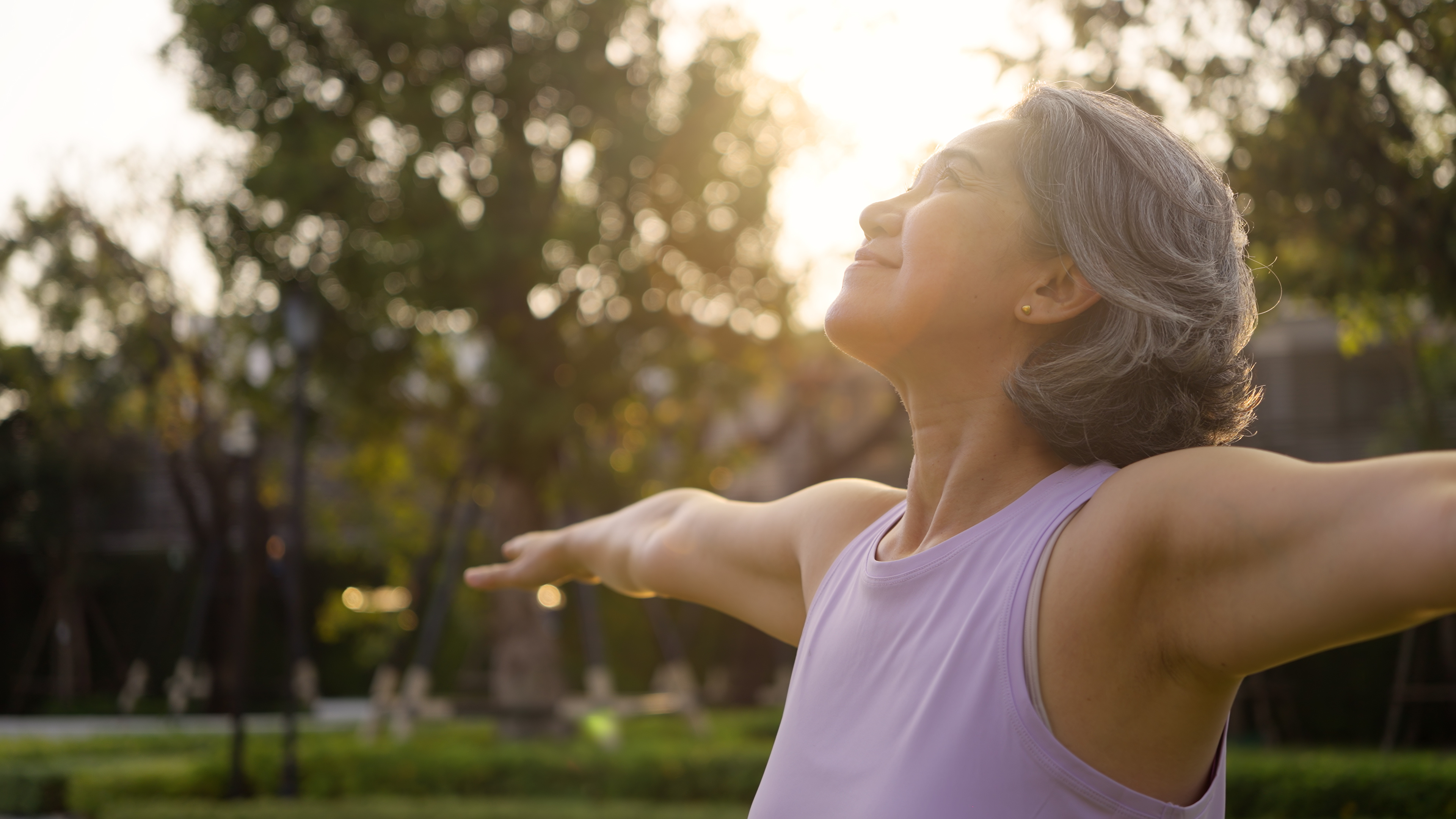 Woman with gray hair enjoying nature, arms outstretched, during sunset.