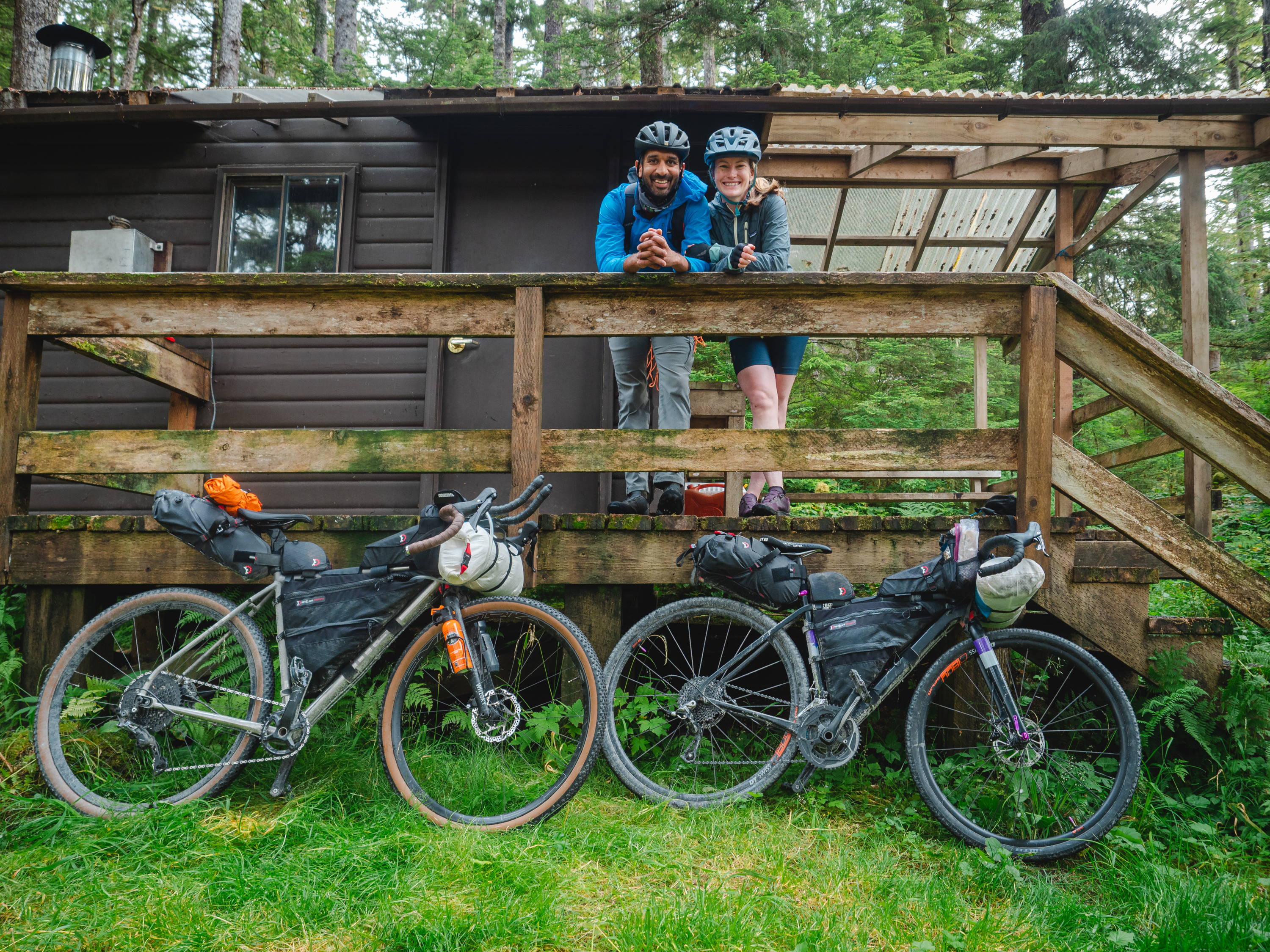 A couple in bikepacking gear stands in front of a woodsy cabin while their Otso bikes lean against the cabin's front railing.