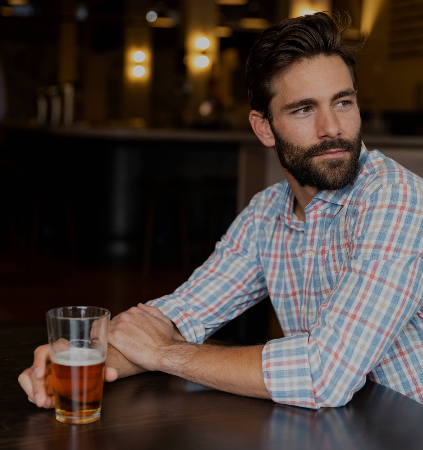 A man sitting in a dark restaurant wearing the casual blue and red Boyne check shirt drinking a beer.