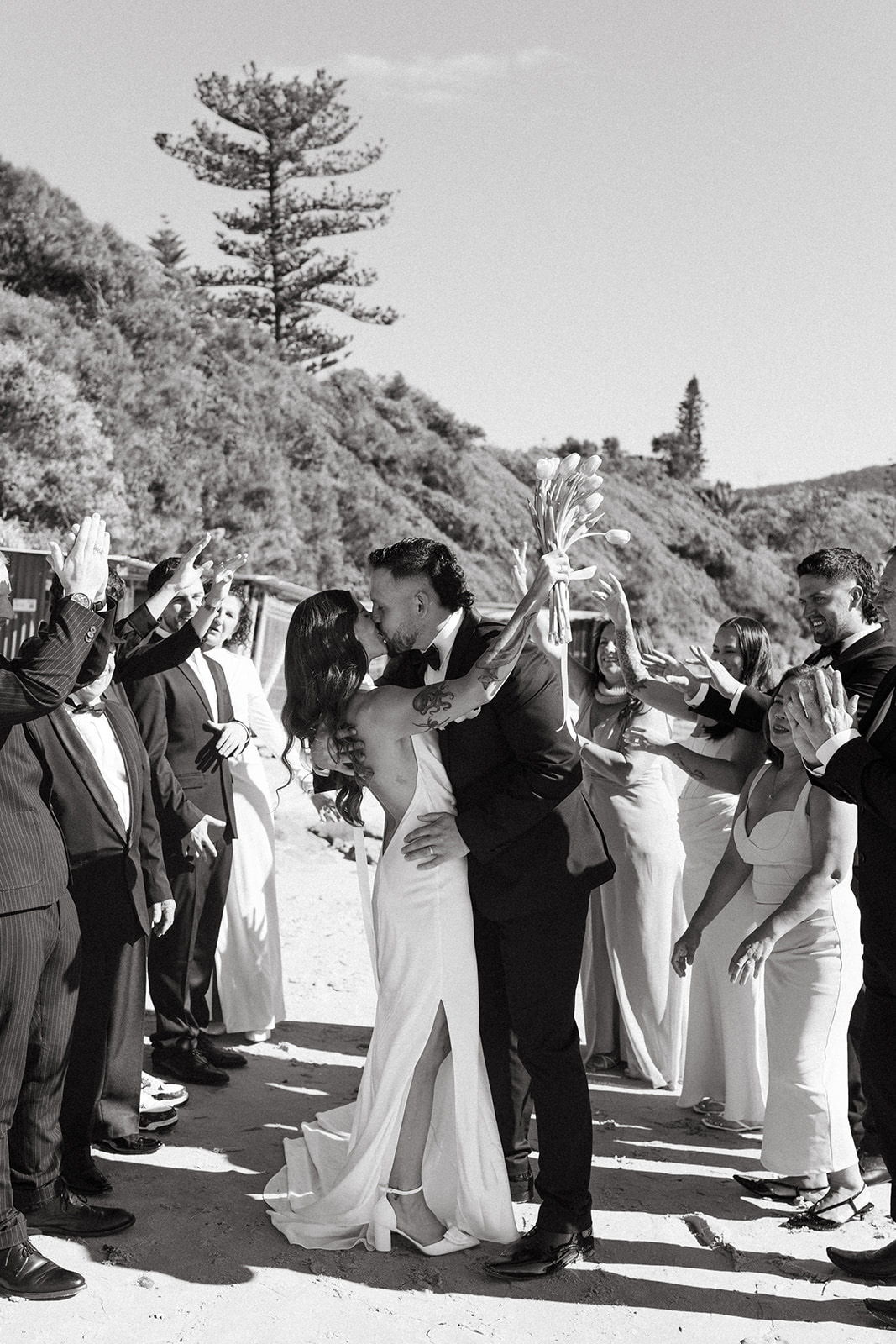 Couple kissing at wedding celebration, surrounded by guests in formal attire.