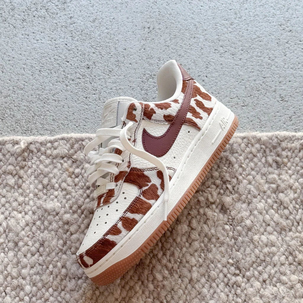 White sneakers with brown cow print and red swoosh on a textured surface.