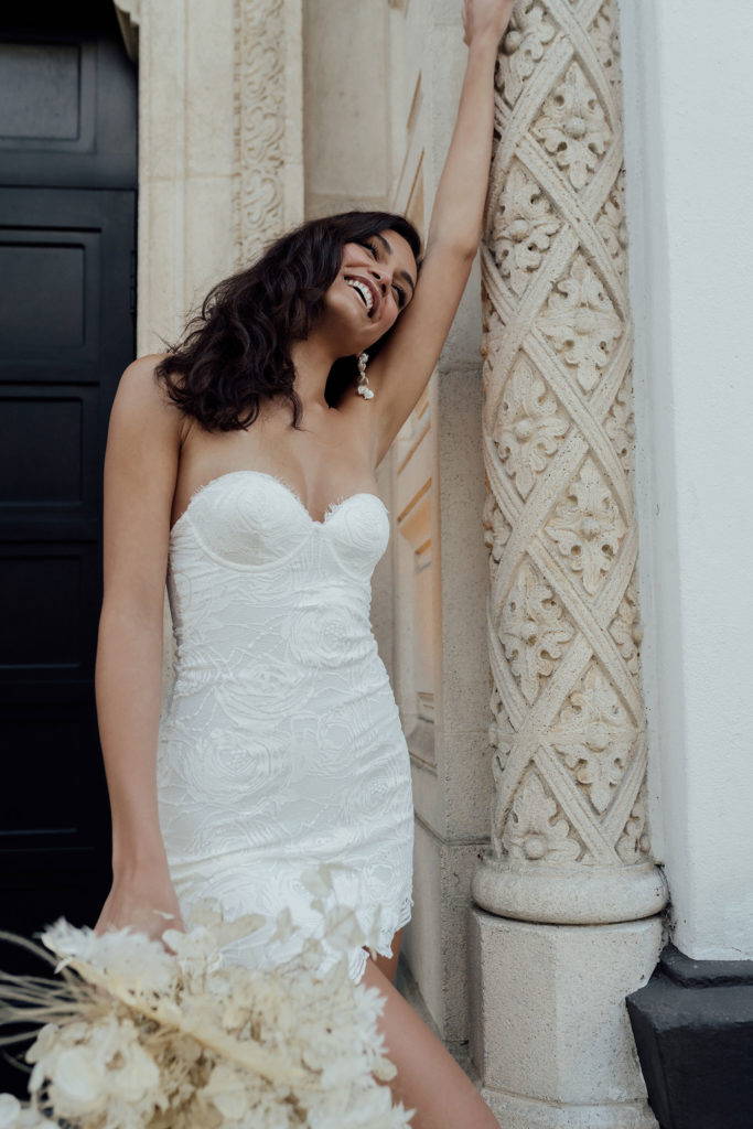 Frau in kurzem Spitzenkleid mit braunem Haar hält Blumenstrauß 