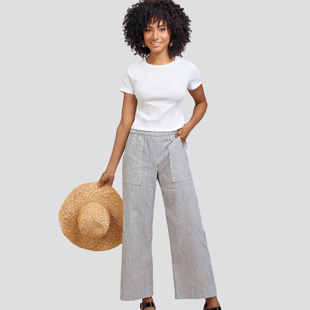 Woman in a white shirt holding a straw hat, wearing wide-leg gray pants.