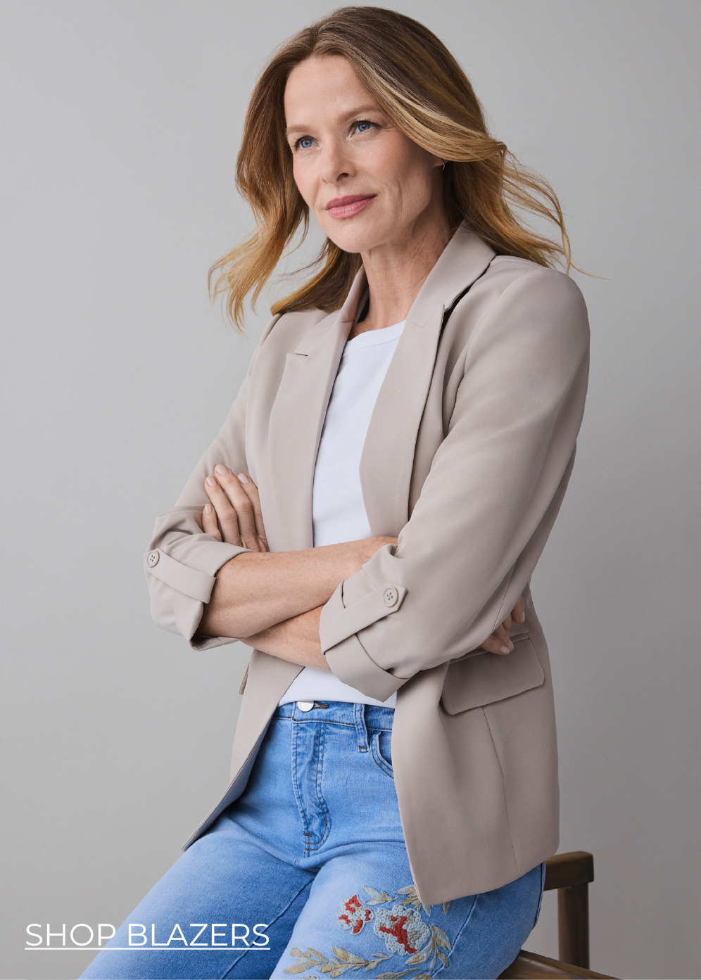 Woman in a beige blazer with arms crossed, smiling against a neutral background.