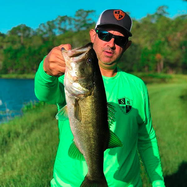 Willie Fernandez at the edge of a lake, holding up a bass fish while wearing a green SA Company cotton long sleeve shirt, sunglasses and a SA Company hat.