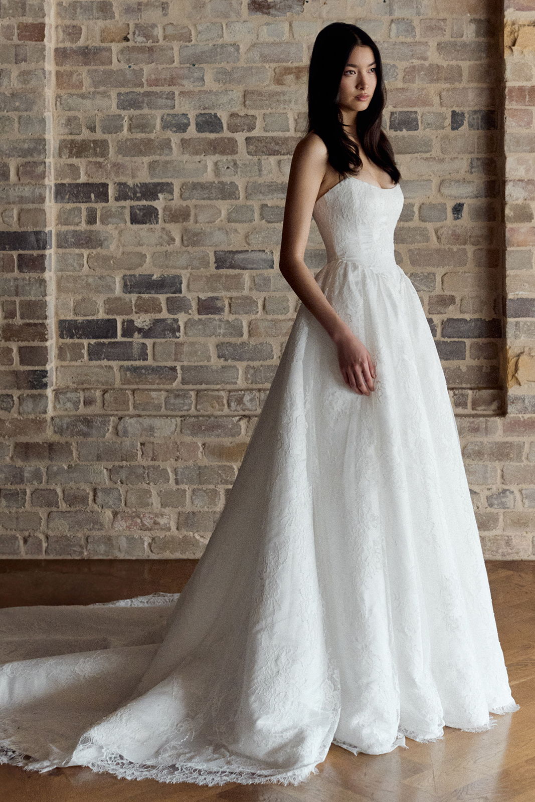A woman in a white wedding dress stands against a brick wall.