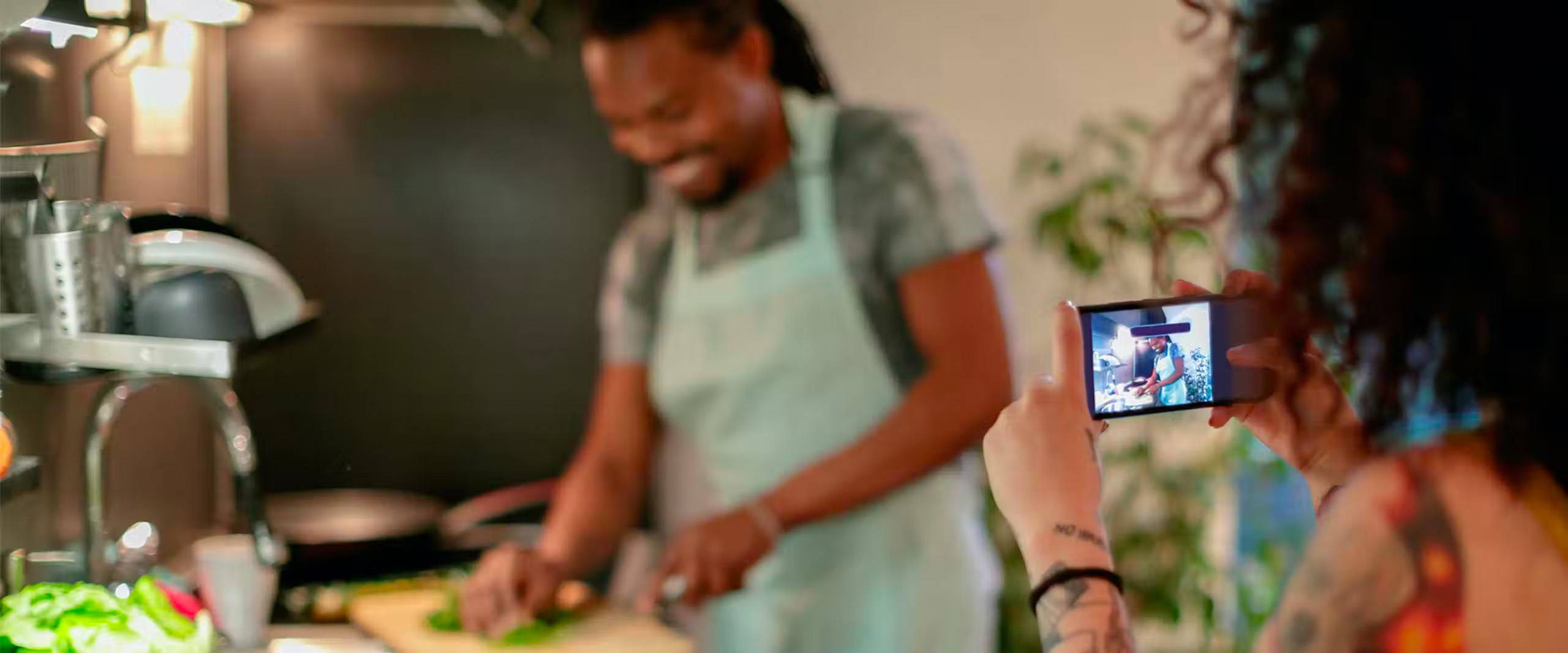 Woman in foreground taking video with her phone of a man, in the background, preparing a meal.