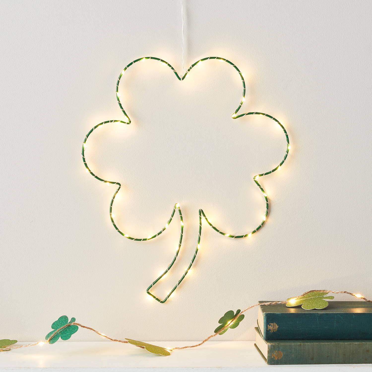 A green fairy light up shamrock decoration hanging on a white wall with a green shamrock garland decoration below.
