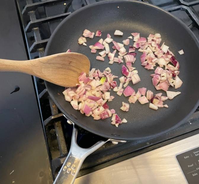 An unseen hand uses a wooden spoon to stir chopped red onion in a Misen Nonstick Pan, which sits on a stovetop.