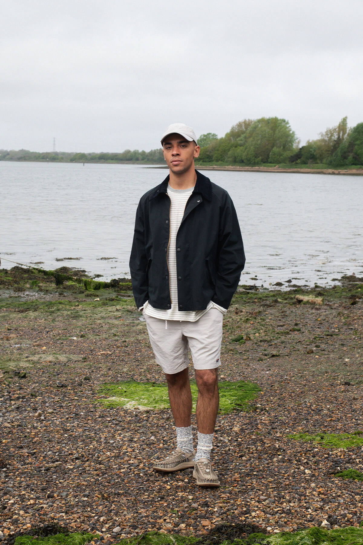 Person standing by a riverbank wearing casual clothing and a cap.