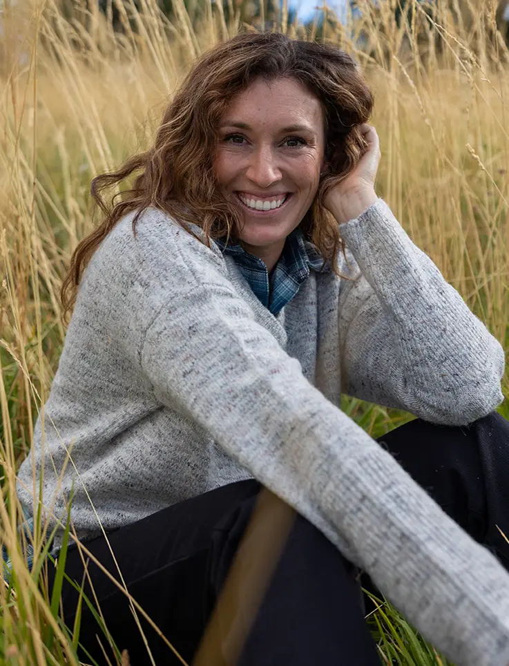 Woman smiling in a field of tall grass, wearing a cozy sweater.